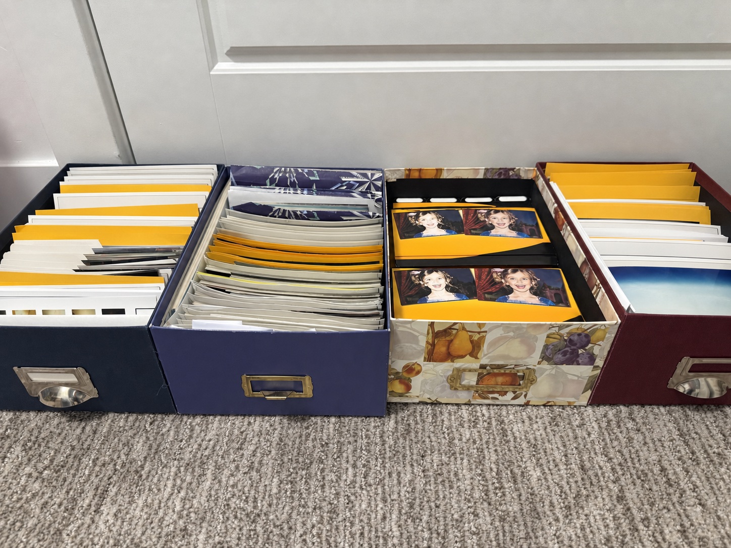 Four open photo storage boxes lined up on the floor, each packed front to back with loose prints and photo lab envelopes